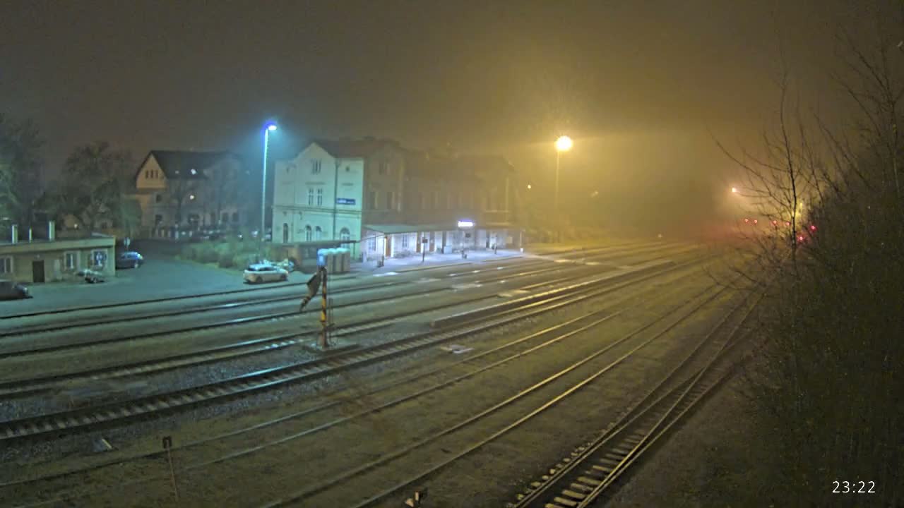A foggy night scene shows a train station with several tracks extending into the distance, illuminated by streetlights.
