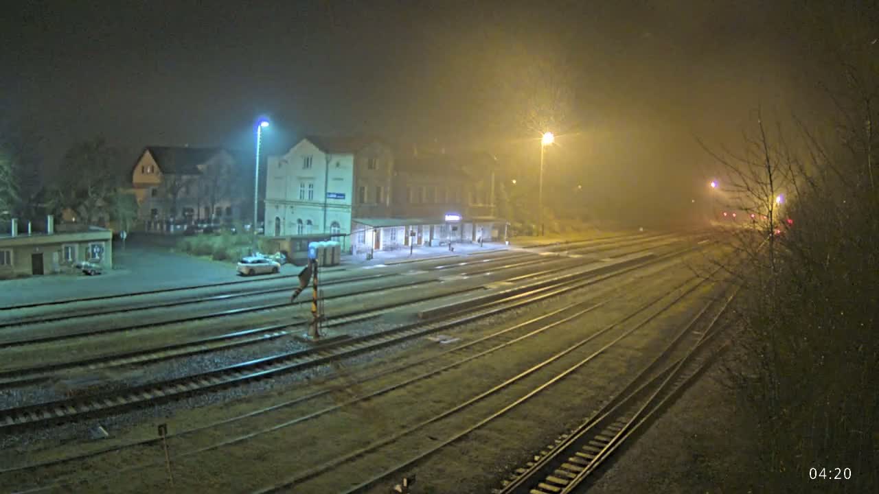 A foggy night scene at a train station with multiple tracks extending into the distance, illuminated by streetlights, showing a building and a few cars in the foreground.