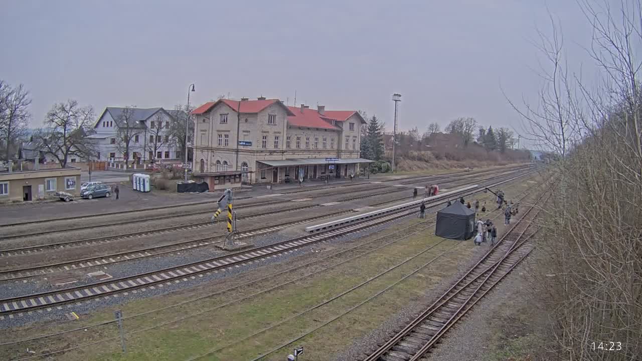 A film crew stands near a train station on a cloudy day next to multiple train tracks.
