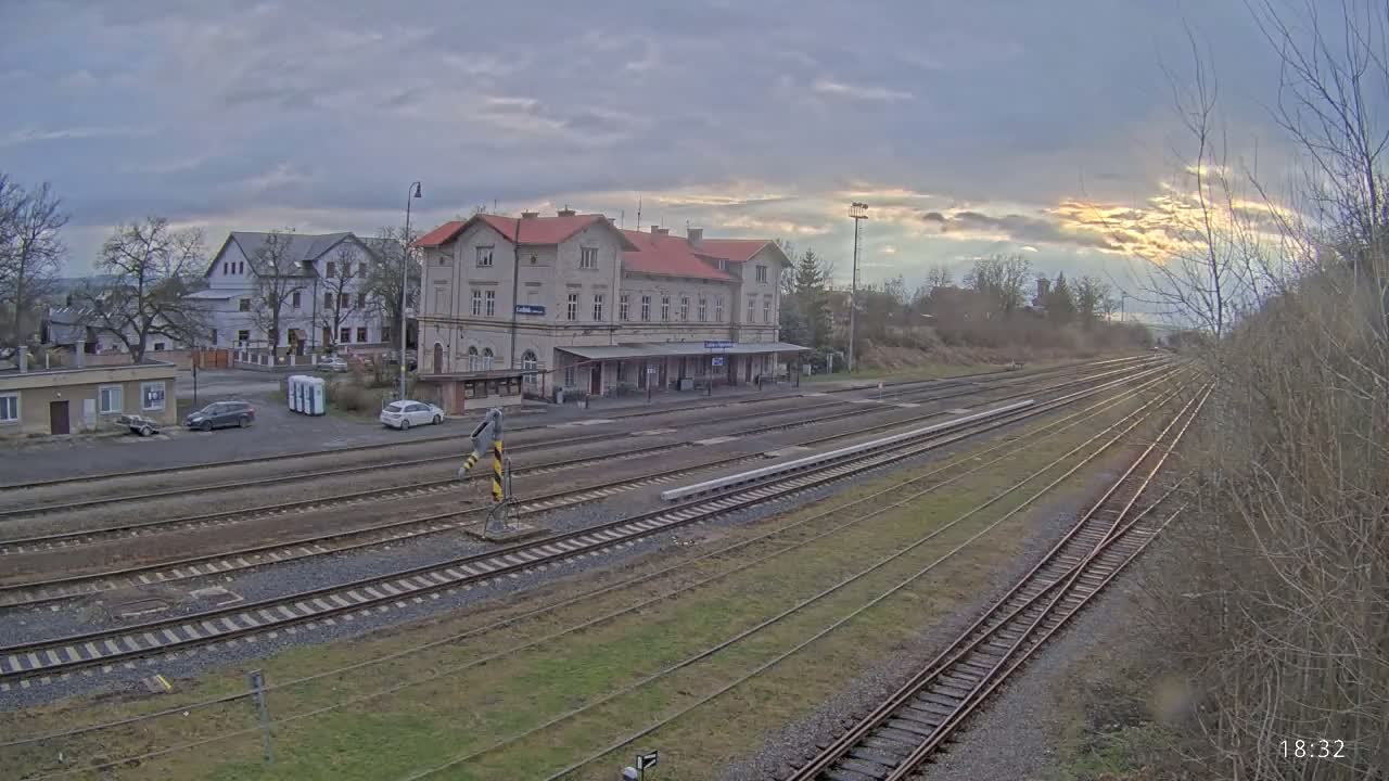 A train station with a long platform and multiple tracks is situated in a town under a cloudy sky with hints of sunset.