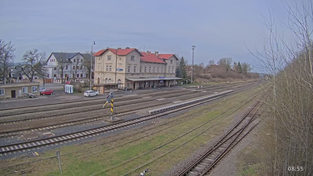 A train station with a long platform and multiple train tracks sits in front of several buildings on an overcast day.