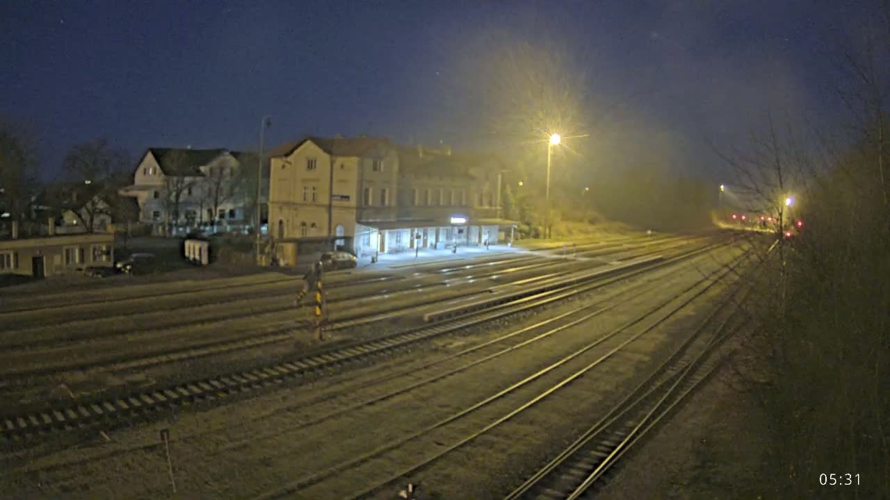 A train station at night, illuminated by artificial light, is situated beside multiple train tracks extending into the foggy distance under a dark sky.