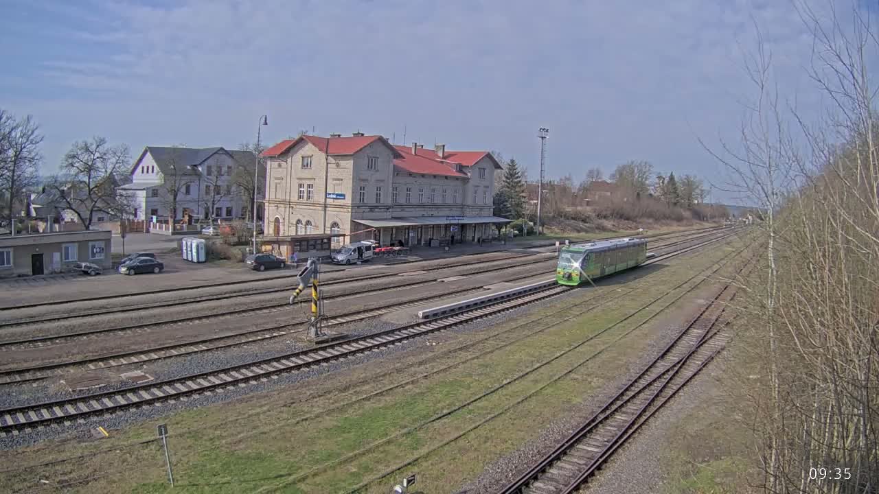 A light-green train is arriving at a train station on a partly sunny day, with a large light-beige building as the main structure.