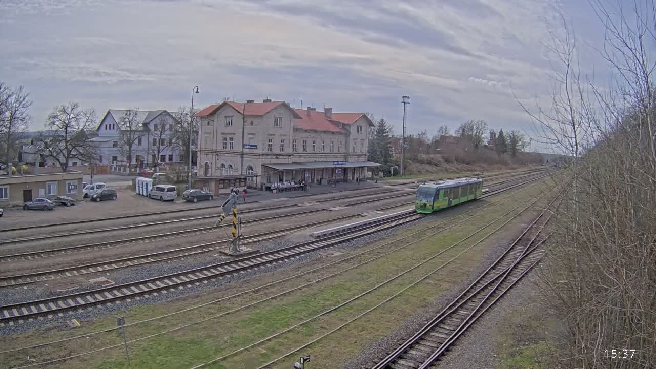A light-green train is at a train station on a cloudy day, with several cars parked nearby and bare trees in the background.