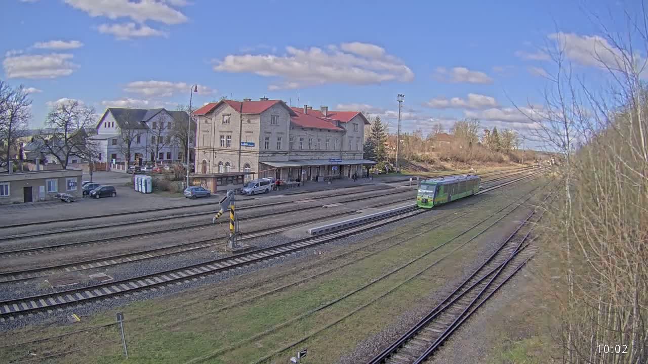 A light-green train sits in a train station on a partly cloudy day, with several tracks and a large station building visible.
