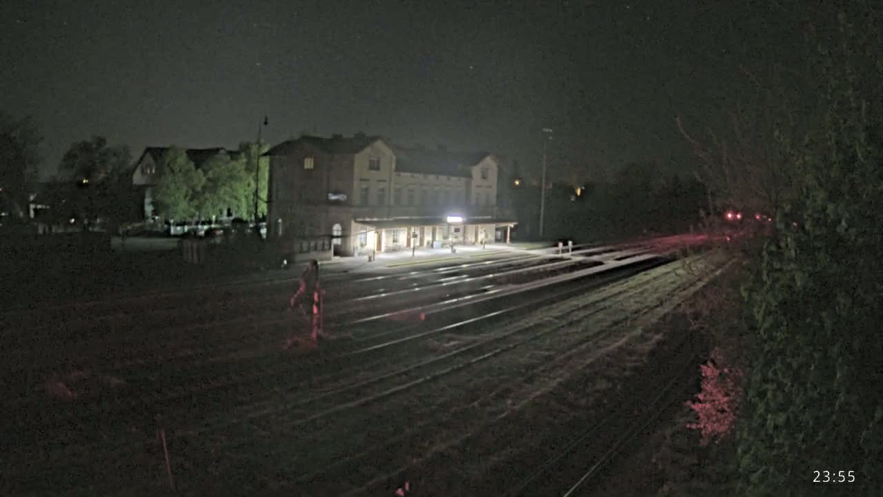 A brightly lit train station at night is situated beside a set of train tracks that extend into the darkness, with some foliage visible near the tracks.