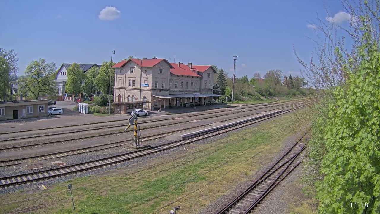 A long view of a train station with a large building, multiple train tracks, and a few cars parked nearby, under a sunny sky with some clouds.
