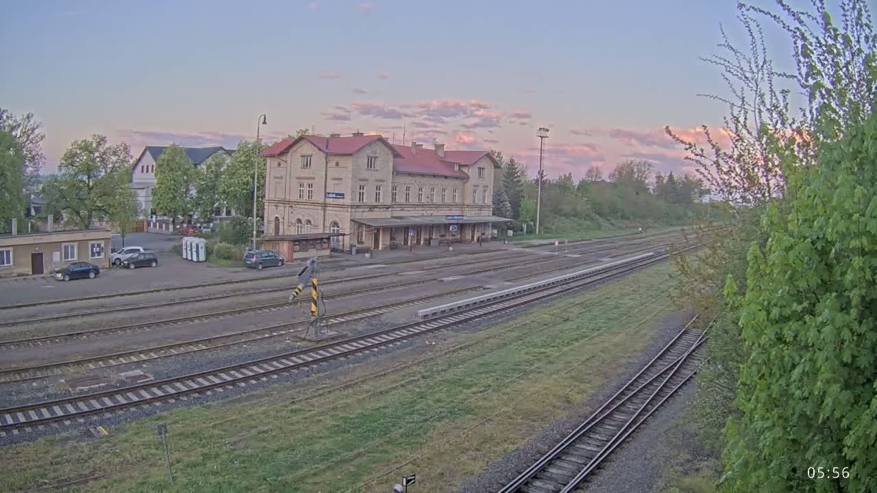A long train station with a few cars parked nearby sits under a pale pink and purple sunrise.