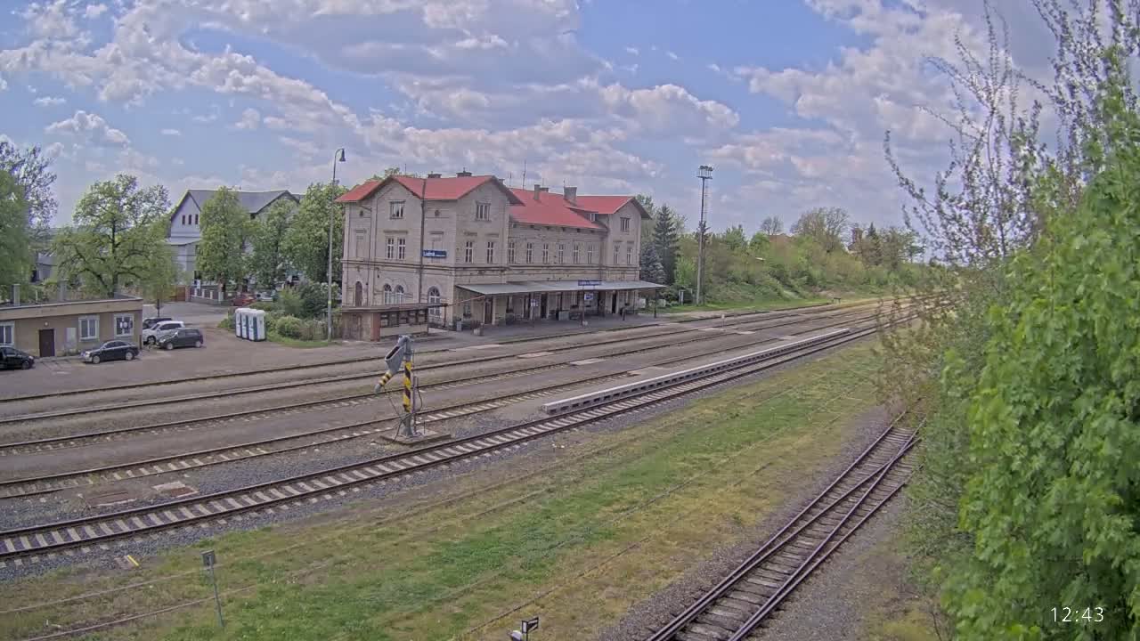 A long shot of a train station with a red-roofed building and several train tracks under a partly cloudy sky.