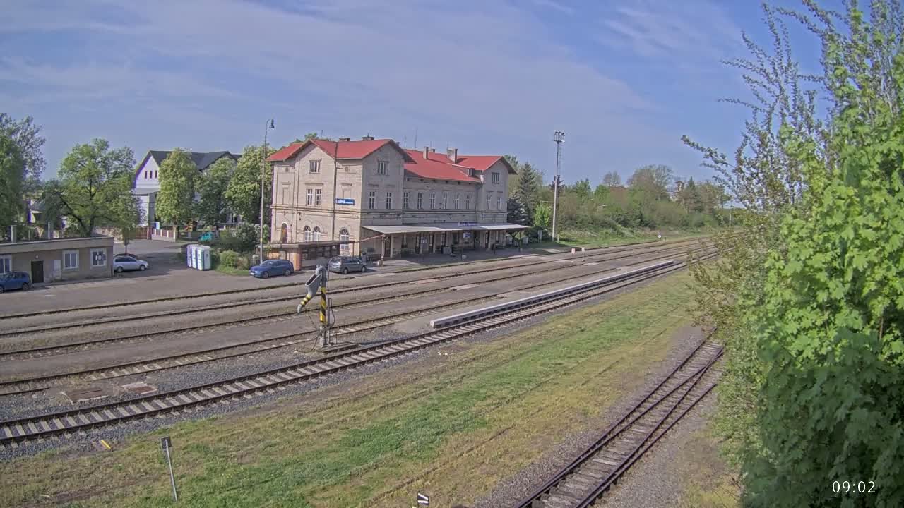 A long shot of a train station on a partly sunny day, with several train tracks leading away from a long, light-colored building with a red roof.