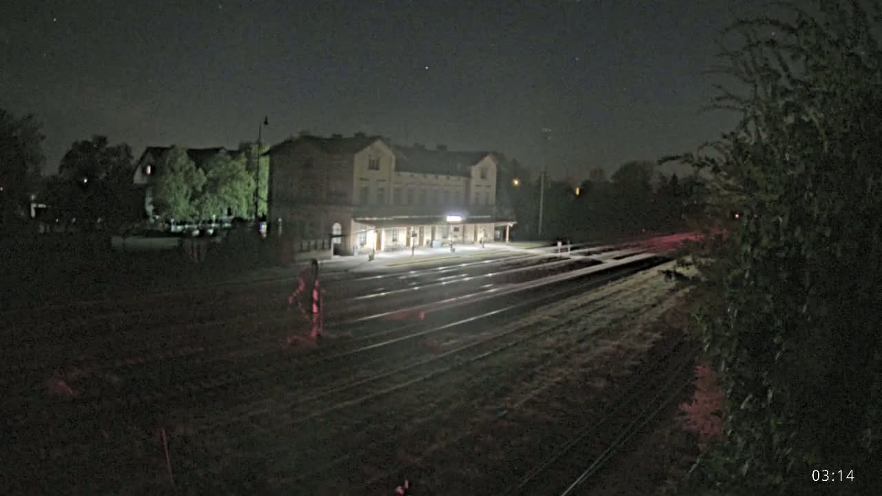A brightly lit train station at night, under a clear, starry sky, is situated beside a set of train tracks.