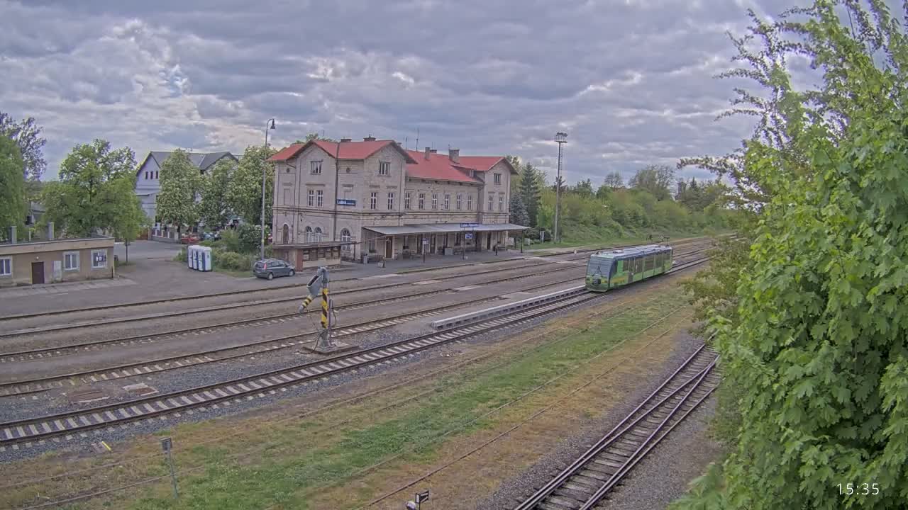 A light-green train approaches a long, pale-yellow train station under a partly cloudy sky.