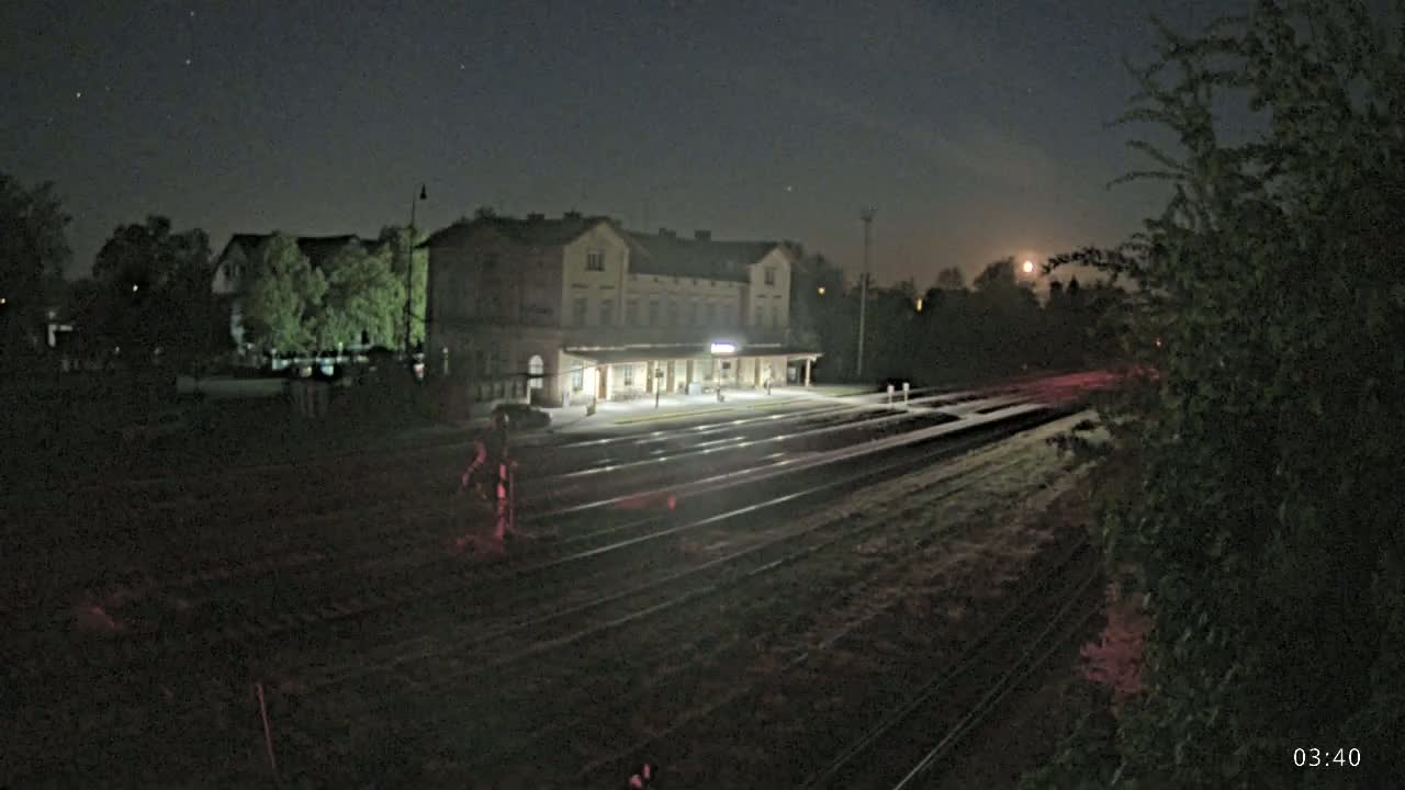 A brightly lit train station is visible under a nighttime sky with a bright moon, surrounded by dark trees and railway tracks.
