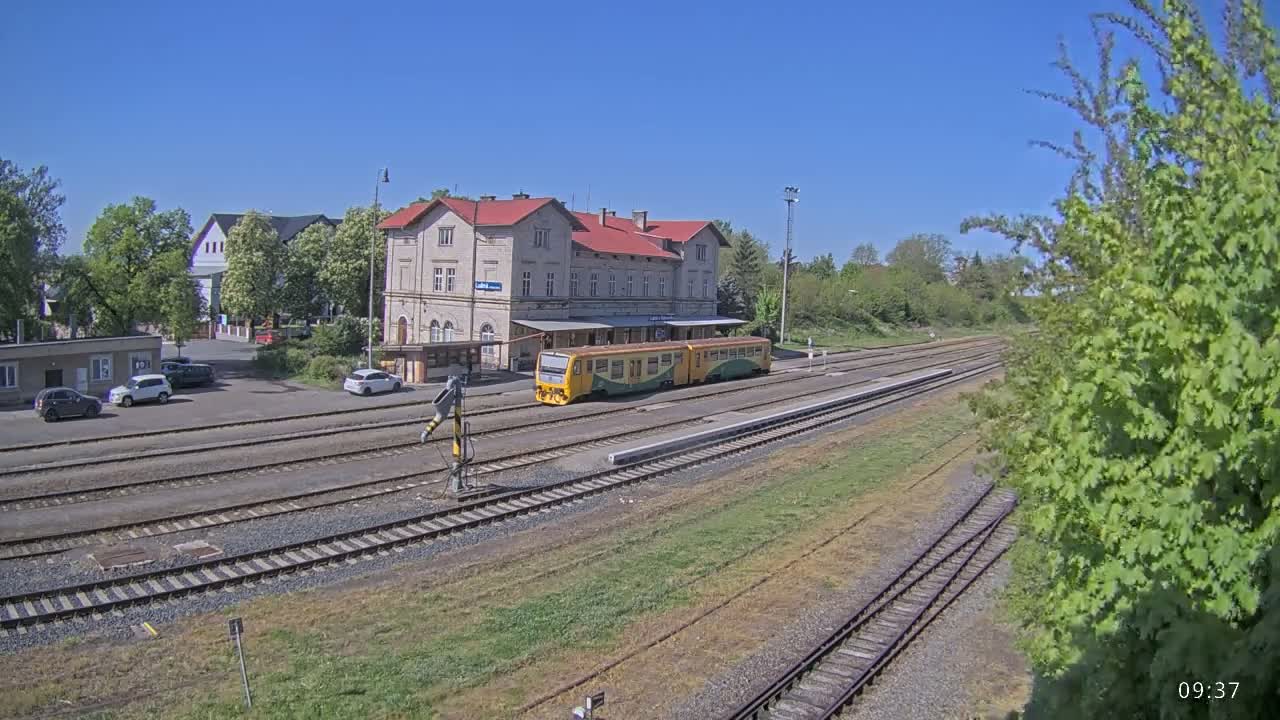 A yellow and green train sits at a train station on a sunny day next to a long building, with a parking lot and several cars in front of it.