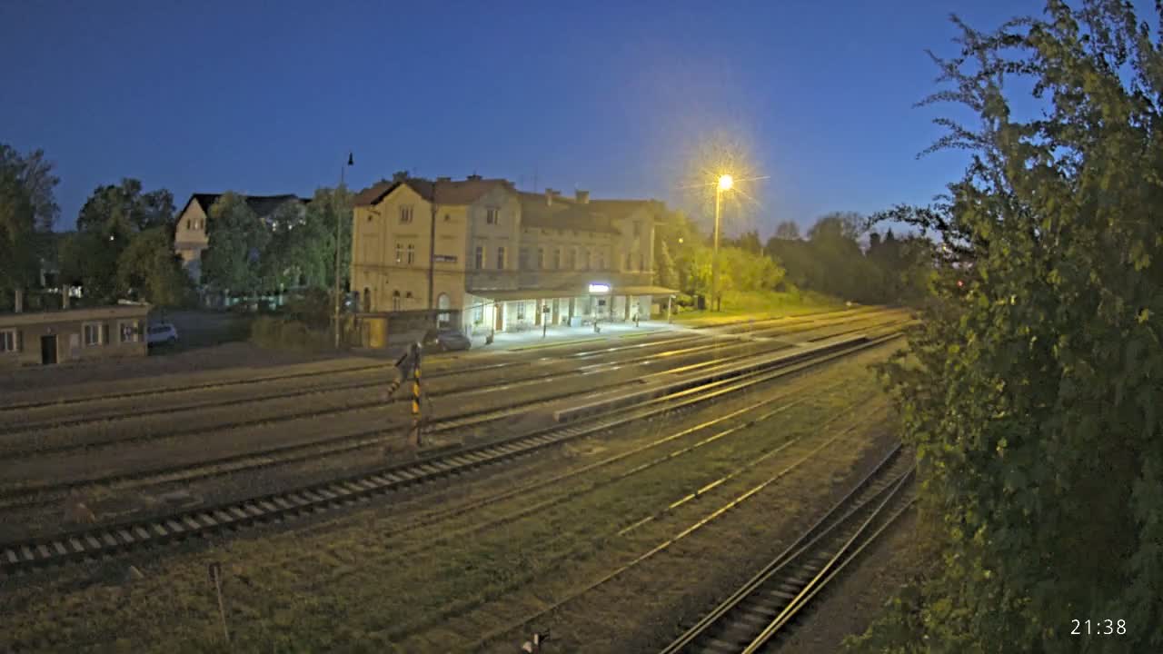 A long view of a train station at night, illuminated by a single lamppost, with multiple train tracks extending into the distance under a clear, dark blue sky.