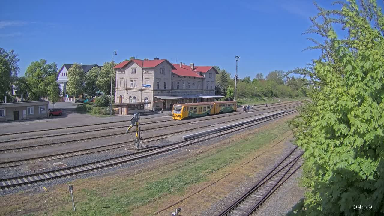 A yellow and green train sits at a station on a sunny day next to a long, red-roofed building.