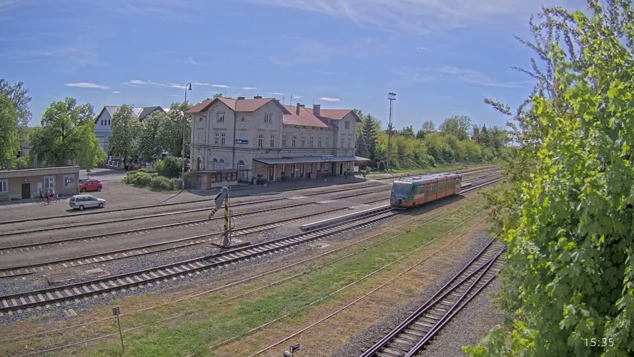 A train is arriving at a long, light-colored train station building on a sunny day.