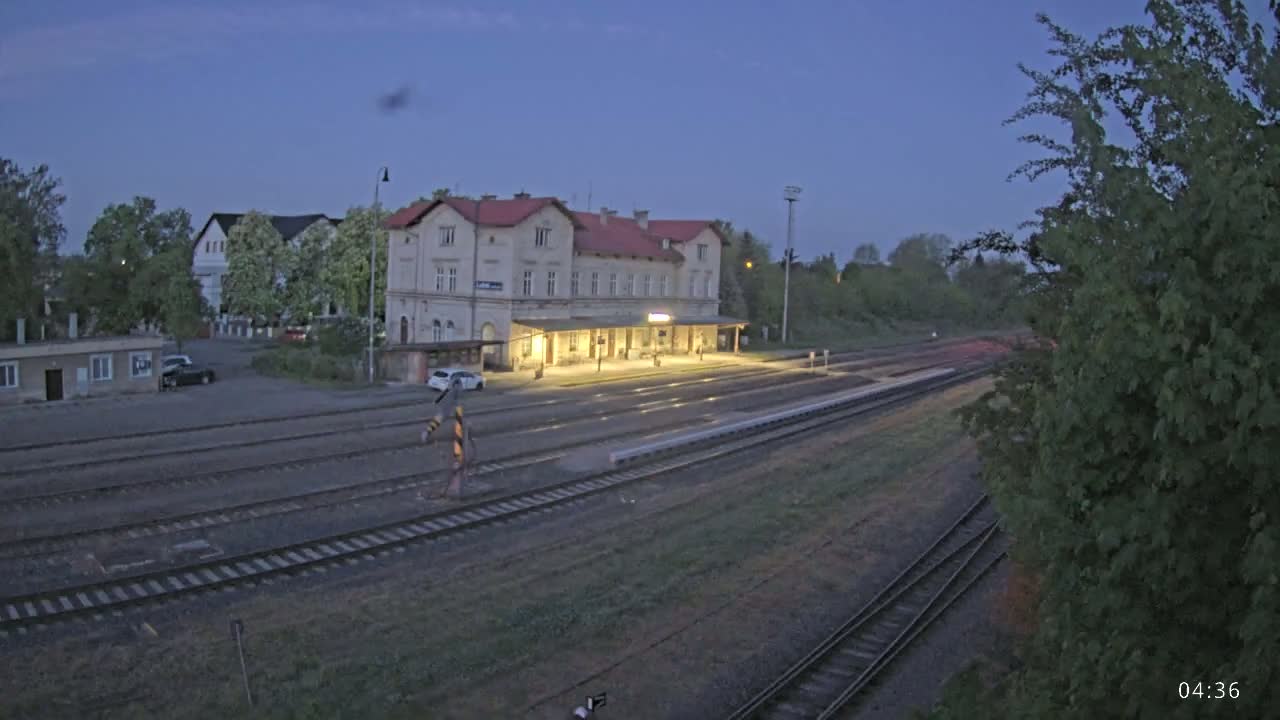 A long, low train station building with a covered platform is situated next to several train tracks under a twilight sky.