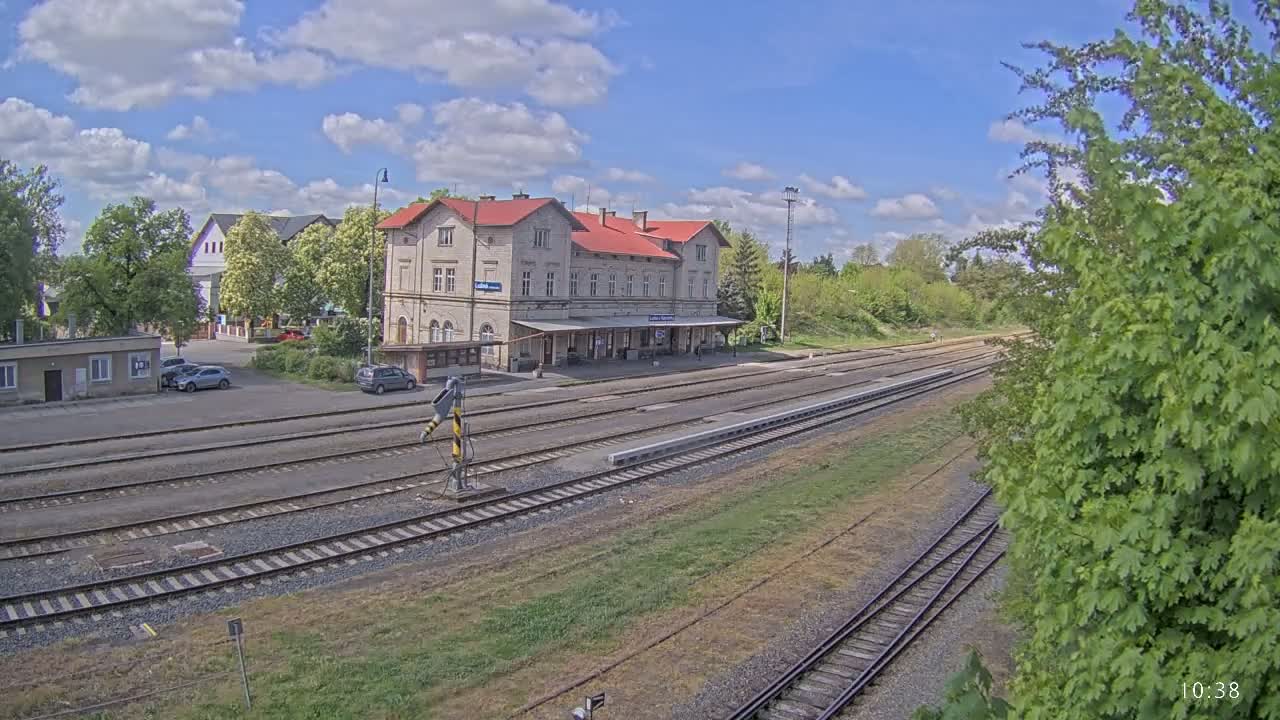A long shot of a train station building with a red roof under a partly cloudy sky, showing multiple train tracks extending into the distance.