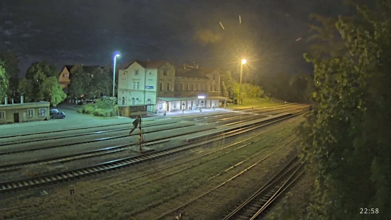 At night, a train station with a long platform and multiple sets of tracks is illuminated by artificial light under a partly cloudy sky.