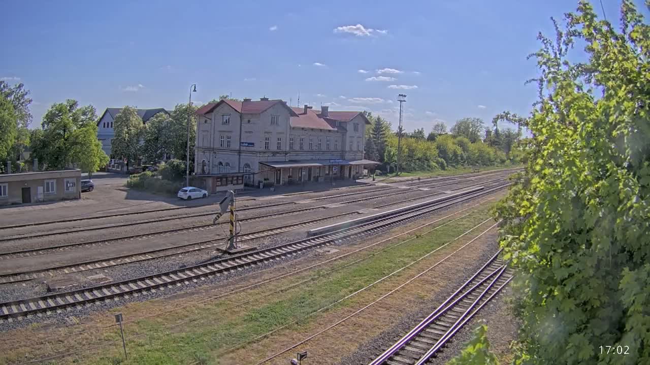 A long, elevated view shows a train station building under a mostly sunny sky with some clouds, surrounded by trees and multiple sets of train tracks.