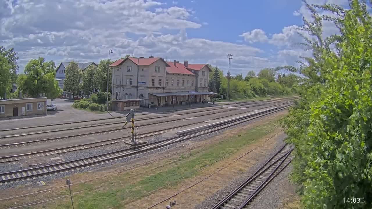 A train station with a long, light-colored building sits beside several train tracks under a partly cloudy sky.