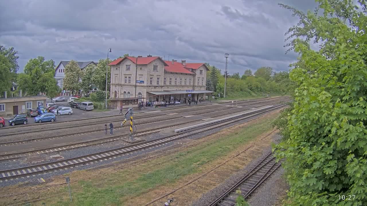A train station with a long platform and several tracks sits under a cloudy sky, surrounded by green trees and parked cars.