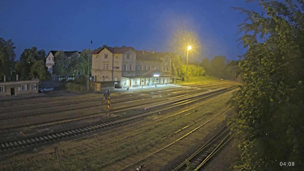 A train station at night, illuminated by a single lamppost, with multiple sets of train tracks extending into the distance under a clear, dark blue sky.