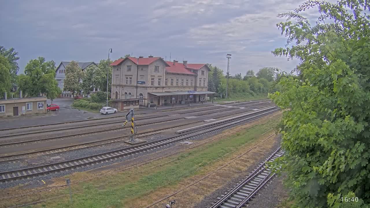 A train station with a long platform and multiple train tracks sits under a cloudy sky, surrounded by green trees and a few parked cars.