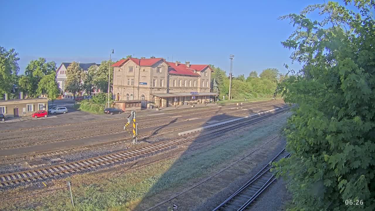A train station with a long building and several tracks sits under a clear, sunny sky.