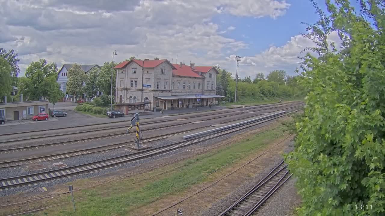 A partly cloudy day reveals a train station with several tracks extending into the distance, surrounded by green trees and a few parked cars.