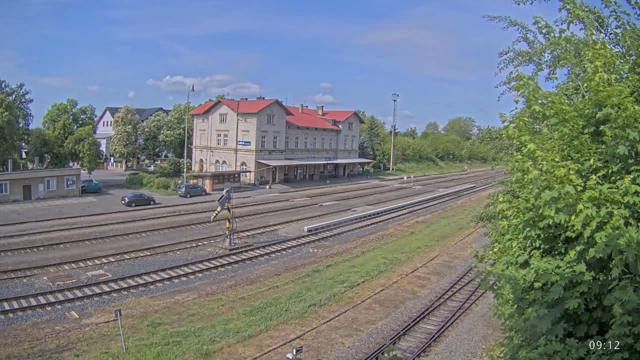 A train station with a long, light-colored building and several train tracks is visible under a mostly sunny sky.