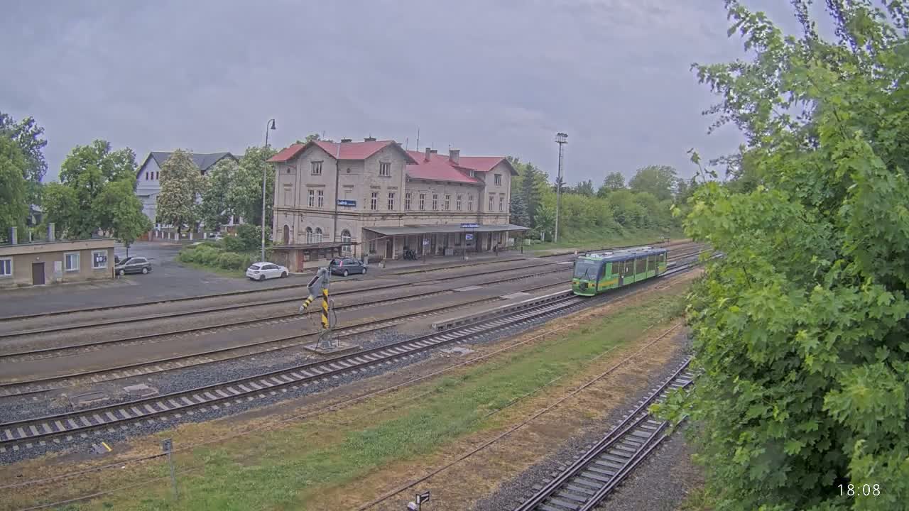 A light-green train is departing a train station on an overcast day, passing by a two-story building with a red roof.