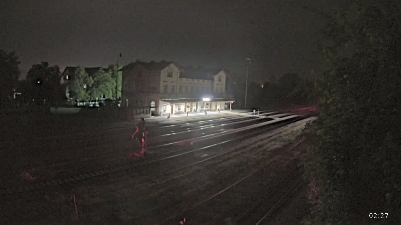 A brightly lit train station at night is seen from across a set of train tracks, under a dark sky.