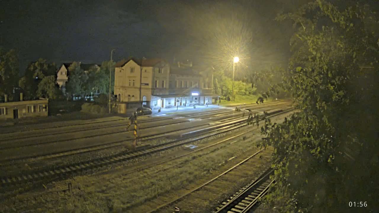 A nighttime view of a train station with multiple tracks, illuminated by streetlights under wet conditions.
