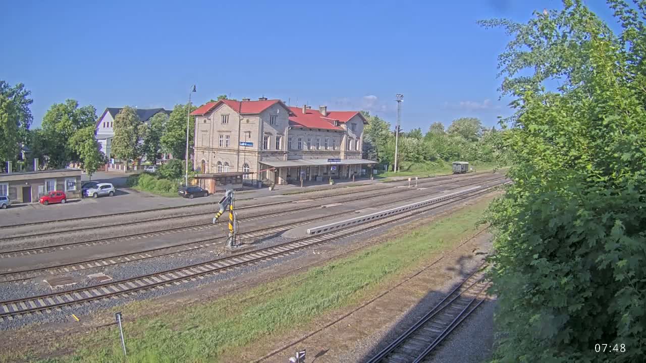 A long train station building with a red roof sits beside multiple train tracks on a sunny day.
