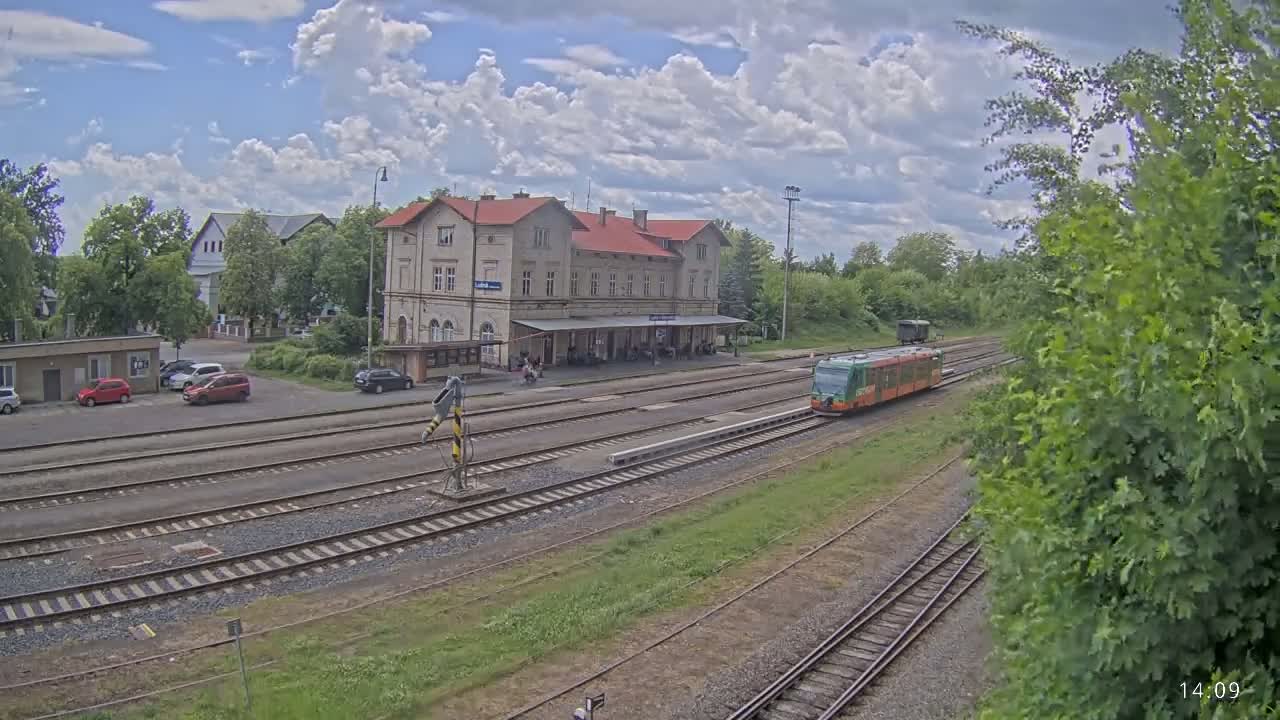 A train pulls into a station with a long building and a parking lot under partly cloudy skies.
