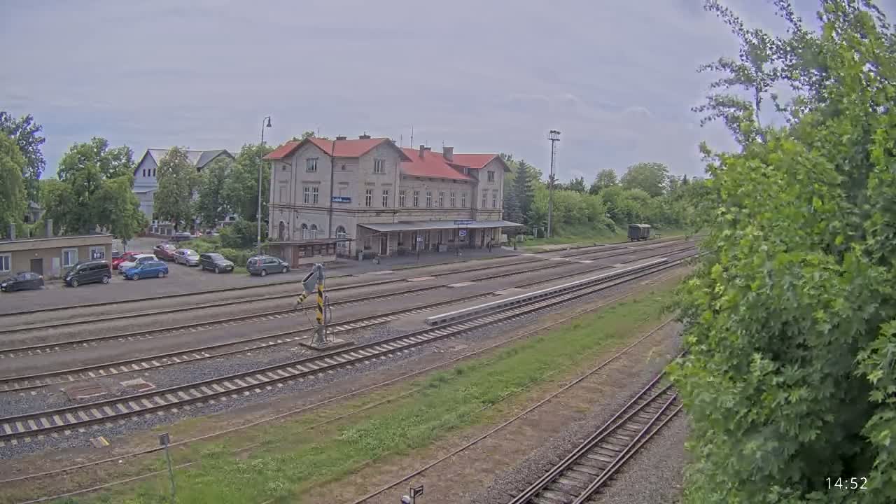 A train station with a long building and several train tracks, under overcast skies, is surrounded by green trees and a parking lot with cars.