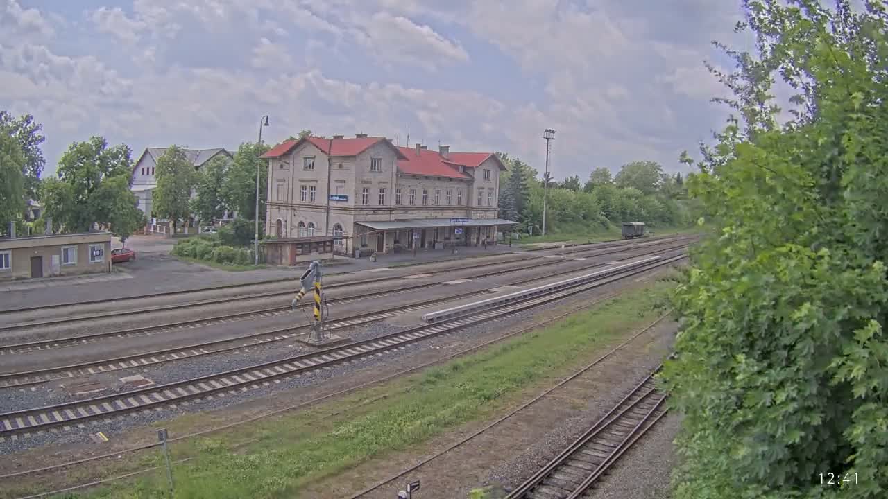 A long view of a train station with a red-roofed building, multiple train tracks, and partly cloudy weather.