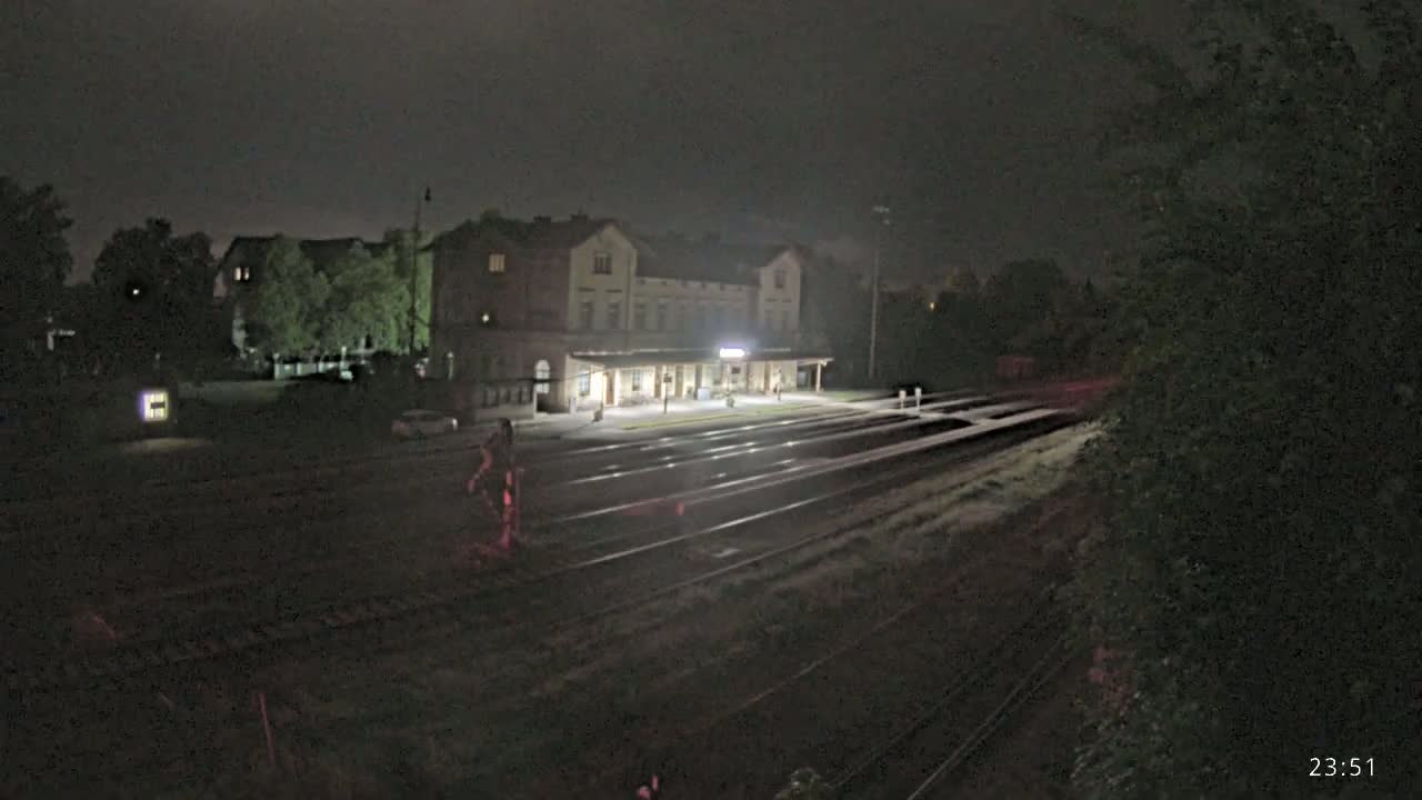 A brightly lit train station at night is visible from across a set of train tracks, under a dark sky.