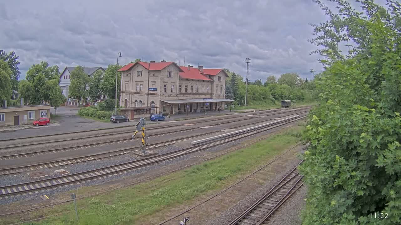 A train station with a long platform and several tracks sits under a cloudy sky, surrounded by green trees and a few parked cars.