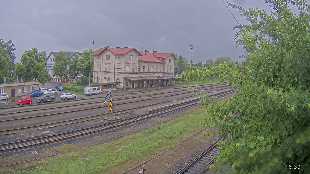 A long, multi-track train station with a large light-colored building and several cars parked nearby under an overcast sky.