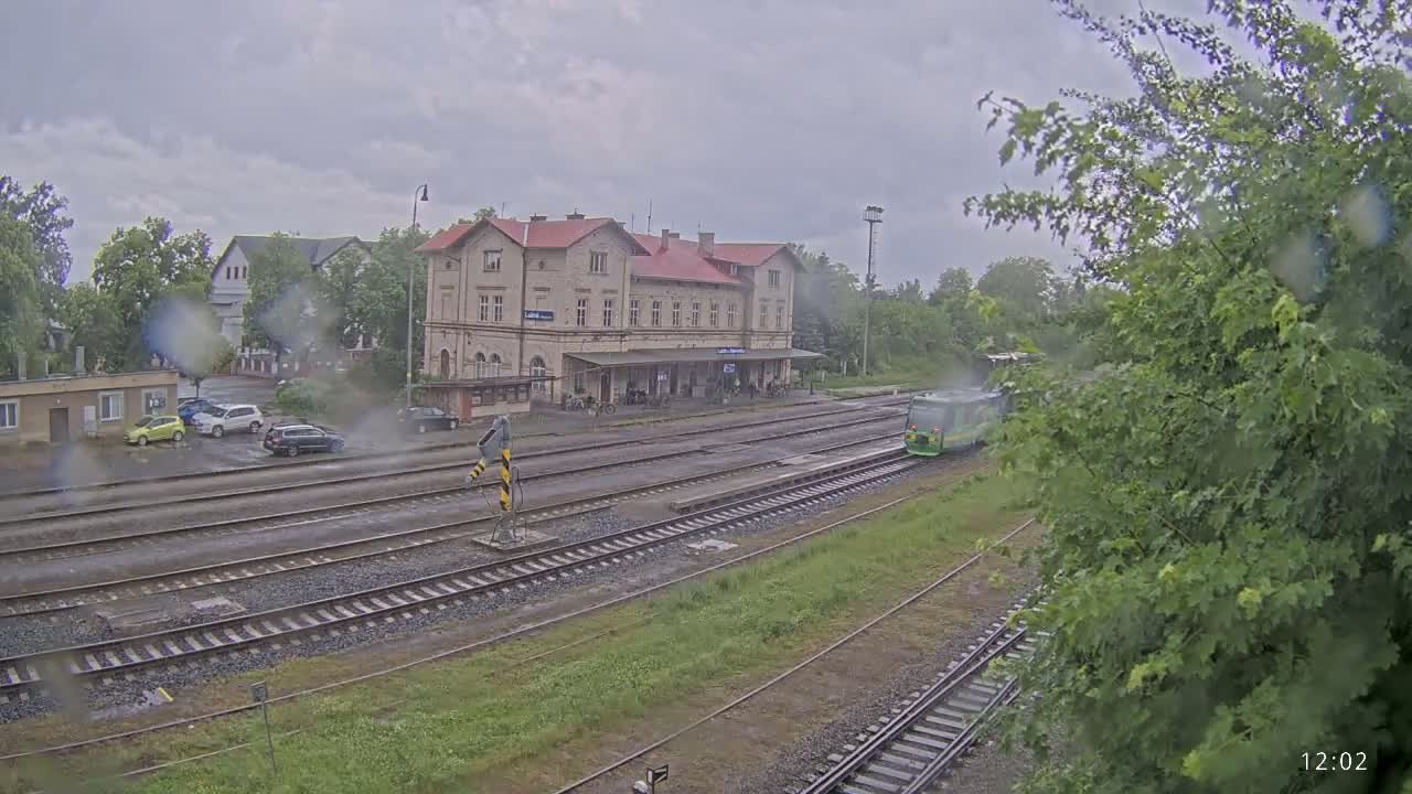A train passes a long, low station building in the rain, with several cars parked nearby.