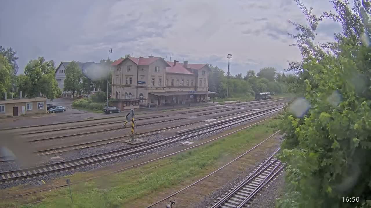 A rain-drenched train station with multiple tracks and a long building sits beside a tree-lined area under a cloudy sky.