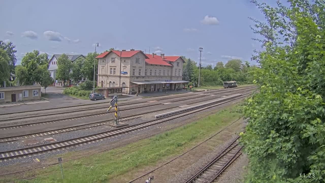 A train station with a long building and several train tracks is visible under a mostly sunny sky.