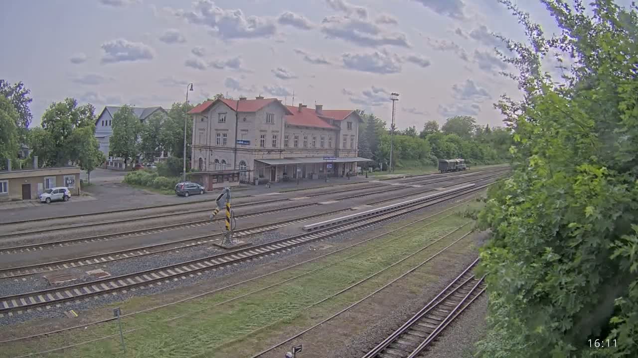 A train station with a long platform and several train tracks is visible under a partly cloudy sky, with a few cars parked nearby and some greenery surrounding the station.