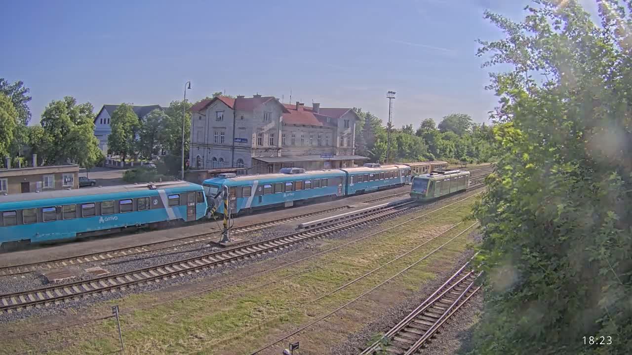 Two light teal passenger trains are at a train station on a sunny day, with one train departing.