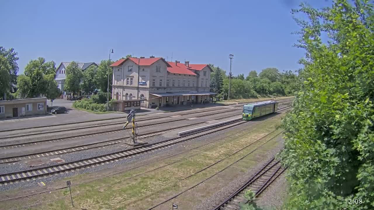 A light-green train sits at a station on a sunny day, with a long building and several tracks visible.