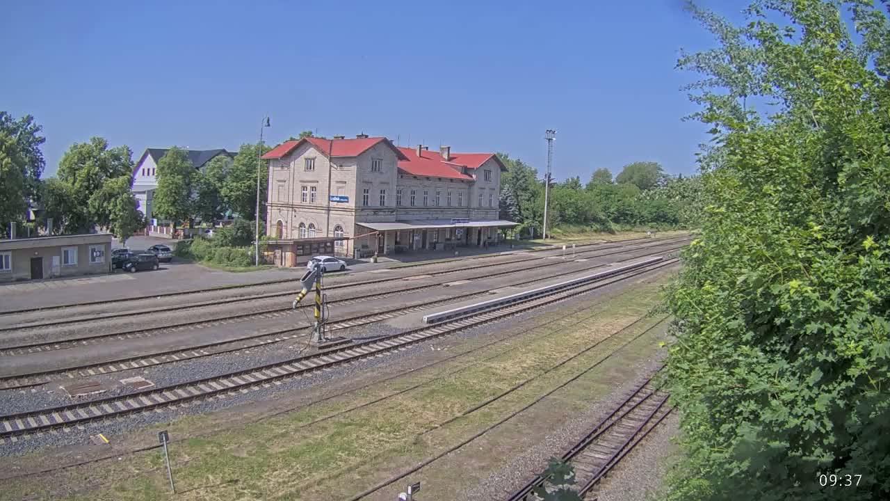 A long, sunny view of a train station with multiple train tracks extending into the distance, surrounded by green vegetation and a few parked cars.