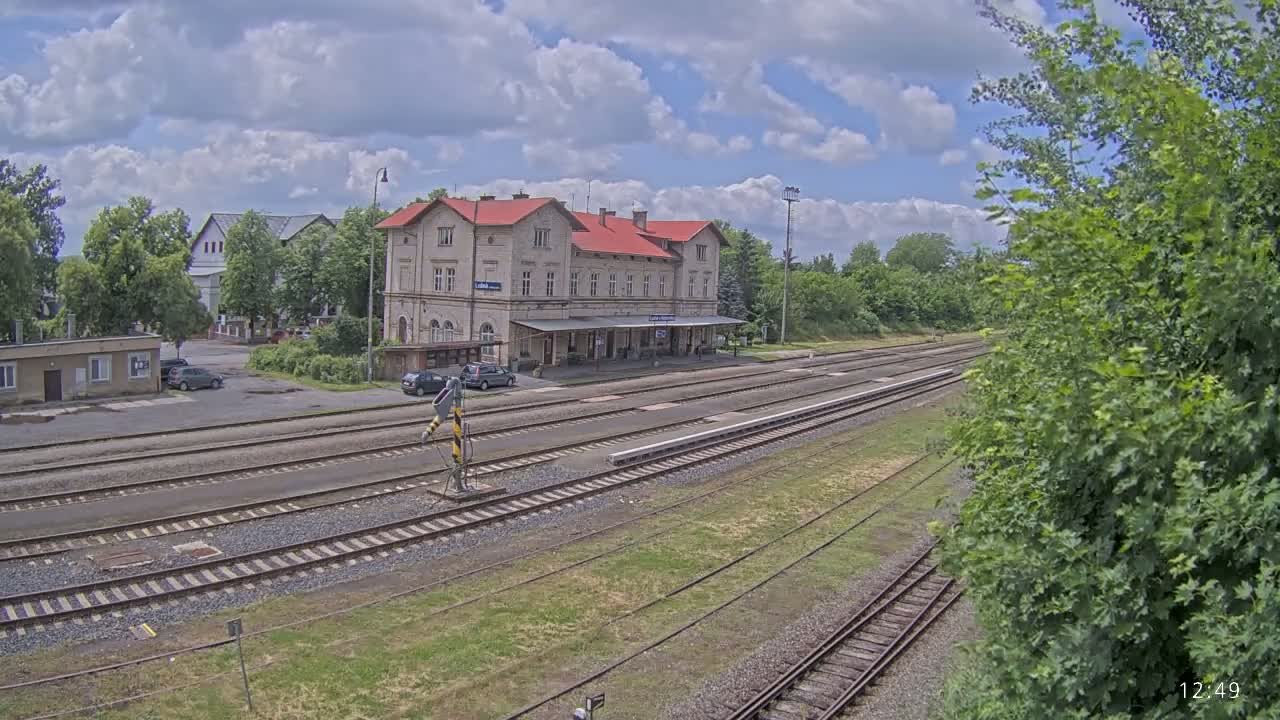 A long shot of a train station with a red-roofed building, several train tracks, and a few cars parked nearby, under partly cloudy skies.